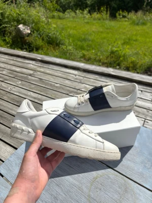Valentino Open Mörkblå - Snygga vita sneakers från Valentno med en bred marinblå läderdetalj över sidan. Skorna har klassisk låg profil, vita snören och en chunky sula med nitar baktill. Perfekta för dig som vill ha en clean men ändå unik look. Allt OG följer även med som visas på sista bilden. Kontakta mig vid frågor, Pris kan även förhandlas vid snabb affär.