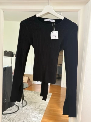 Svart ribbad topp från Bershka - Trendig svart ribbad topp från Bershka med långa ärmar och snygg knytning i ryggen. Toppen har en figurnära passform och slitsade ärmslut för en cool look. Tröjan är i storlek L men passar även någon som bär S. 