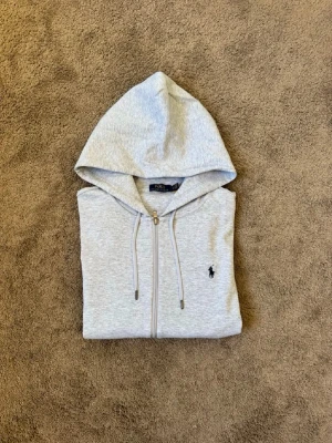 Grå zip hoodie från Polo Ralph Lauren - Snygg grå hoodie med dragkedja från Polo Ralph Lauren. Klassisk design med huva och dragsnören, två fickor framtill och den ikoniska mörkblå polospelaren broderad på bröstet. Tillverkad i mjuk bomullsblandning för skön känsla.