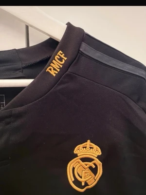 Real Madrid svart matchtröja Adidas - Snygg svart Real Madrid matchtröja från Adidas med gula detaljer och klubbmärke på bröstet. Tröjan har korta ärmar, rund hals och texten 'Emirates Fly Better' framtill. Perfekt för fotbollsträning eller att bära till vardags. Xxl