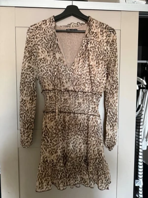 Leopardmönstrad kortklänning från Zara - Säljer en snygg leopardmönstrad kortklänning från Zara med v-ringning, långa ärmar och smock i midjan. Klänningen har volangkant nedtill och är tillverkad i ett lätt, luftigt material som passar perfekt för en trendig look.
