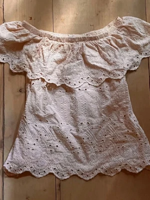 Vit offshoulder blus med spetsdetaljer - Söt vit offshoulder blus med broderade spetsdetaljer och vågig kant. Blusen har korta ärmar och ett romantiskt mönster med hålbroderier. Perfekt för varma dagar och ger en bohemisk vibe.