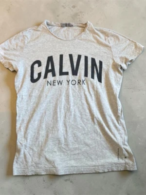 Grå Calvin Klein t-shirt - Snygg grå t-shirt från Calvin Klein med stort svart tryck 'CALVIN NEW YORK' på bröstet. Klassisk rund halsringning och korta ärmar. Tillverkad i mjuk bomull för en bekväm känsla. Perfekt till jeans eller shorts. Bra skick, i storlek s. Passar för kille och tjej, men främst för tjejer