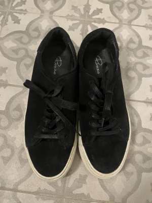 Svarta sneakers från Rizzo - Stilrena svarta sneakers från Rizzo i mocka med vita sulor. Klassisk låg modell med svarta skosnören och diskret logga på insidan. Perfekta för dig som gillar enkel och tidlös design.