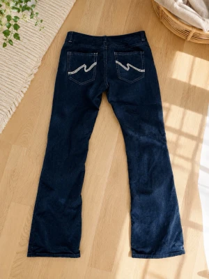 Mörkblå bootcut jeans med broderi - Snygga mörkblå bootcut jeans med vita broderade detaljer på bakfickorna. Klassisk femficksmodell med knapp och dragkedja framtill. Jeansen har en lätt utsvängd passform och är tillverkade i ett mjukt denimtyg.