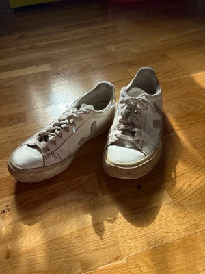 Vita Veja sneakers i läder - Säljer ett par klassiska vita Veja sneakers i lågt snitt. Skorna är tillverkade i läder med den ikoniska V-loggan på sidan och har vita skosnören samt en beige sula. Perfekta för en clean och stilren look.