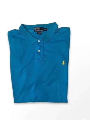 Blå pikétröja från Ralph Lauren - Klassisk blå pikétröja från Ralph Lauren med korta ärmar och krage. Tröjan har två knappar framtill och den ikoniska gula logotypen broderad på bröstet. Tillverkad i mjuk bomull för en bekväm känsla.