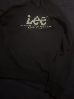 Mörkblå hoodie från Lee med tryck - Säljer en mörkblå hoodie från Lee med stort vitt tryck på bröstet där det står 'Lee Crafted With Purpose Since 1889'. Klassisk modell med huva och dragsko. Perfekt för dig som gillar streetwear och vill ha en enkel men snygg hoodie.