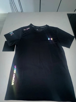 Svart Under Armour t-shirt - Snygg svart t-shirt från Under Armour med regnbågsfärgat logotryck på bröstet och text längs sidan. T-shirten har korta ärmar och rund hals. Perfekt för dig som gillar sportig och modern stil.