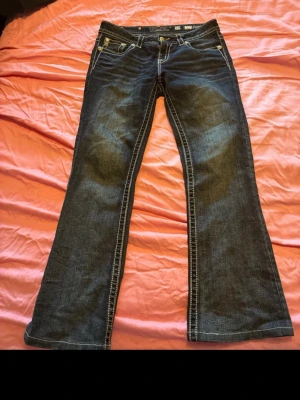 Miss me jeans  - Säljer ett par mörkblå bootcut jeans i mid rise från Miss me med snygga broderade bakfickor i guld och lila. Jeansen har kontrastsömmar  och dekorativa nitar. Perfekta för dig som vill sticka ut lite extra med detaljerad design. Säljer även på vinted och pris kan alltid diskuteras det är ett dyrt märke 💕