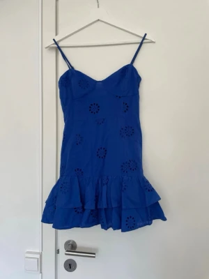  Trendig Blå kort klänning med volang - Snygg blå kort klänning med smala axelband och volangkant nertill. Klänningen har broderade blommönster och en omlottliknande byst. Perfekt för varma dagar och sommarens alla tillfällen.