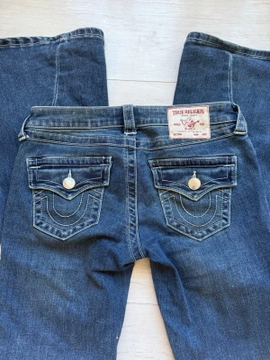 Blå bootcut jeans från True Religion - Säljer ett par blå bootcut jeans från True Religion med ikoniska ficklock och vita kontrastsömmar. Jeansen har låg midja, klassiska silverknappar och är tillverkade i denim. Perfekta för dig som gillar en avslappnad men snygg stil. 