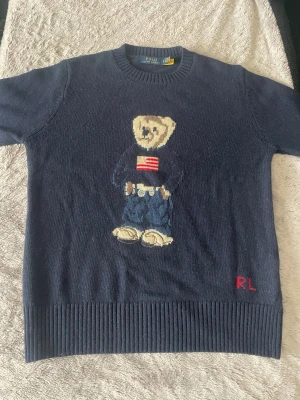 Mörkblå stickad tröja Polo Bear - Mörkblå stickad tröja från Ralph Lauren med den ikoniska Polo Bear på bröstet, klädd i jeans och tröja med amerikansk flagga. RL-broderi i rött nedtill. Rund halsringning och ribbade muddar. Perfekt för dig som gillar klassisk preppy stil. Storlek M kan passa S