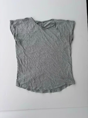 Grå basic t-shirt i bomull - En enkel grå t-shirt i mjuk bomull med korta ärmar. Perfekt till vardags och lätt att matcha med olika stilar. T-shirten har en avslappnad passform och är helt utan mönster eller tryck. Storlek 146/152. Ett väldigt litet hål vi armhålan (syns knappt).
