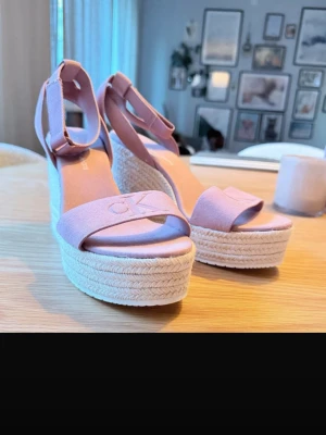 Rosa kilklackade sandaler Calvin Klein - Säljer ett par ljusrosa sandaler från Calvin Klein med hög kilklack i flätad jute. Sandalerna har öppen tå, bred rem med CK-logga framtill och justerbart ankelband. Perfekta för sommaren och ger extra höjd med sin trendiga espadrille-look.