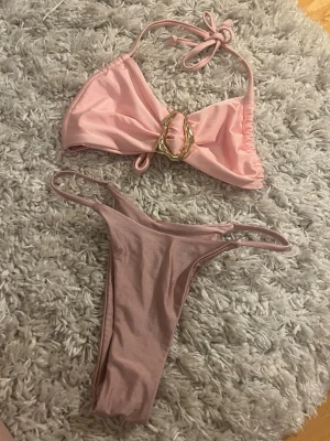 Ljusrosa bikini med gulddetalj - Trendig ljusrosa bikini med bandeau-topp som har en snygg guldfärgad detalj i mitten och knytband i nacken. Matchande bikiniunderdel med smala band i sidorna. Perfekt för stranden eller poolen!