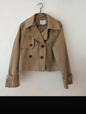 Kort beige trenchcoat med knappar - Snygg kort trenchcoat i beige med dubbelknäppning och stora svarta knappar. Jackan har bred krage, långa ärmar med detaljer vid ärmsluten och en loose passform. Perfekt för dig som vill ha en trendig och stilren look.