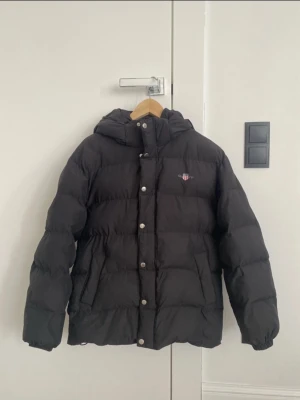 Svart pufferjacka från Gant - Säljer en svart pufferjacka från Gant med huva och dragkedja samt tryckknappar framtill. Jackan har två sidofickor och en liten Gant-logga på bröstet. Perfekt för kalla dagar och riktigt skön att bära tack vare det vadderade materialet.
