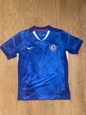 Chelsea FC matchtröja Nike blå - Chelsea FC matchtröja i blått med geometriskt mönster, officiellt klubbmärke på bröstet och vit Nike-logga. Tröjan har korta ärmar, V-ringning med vit och röd detalj samt vita paneler på sidorna. Tillverkad i lätt och ventilerande polyester.