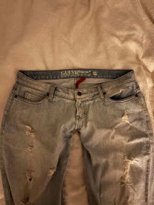 Ljusblå slitna jeans från Guess - Säljer ett par ljusblå jeans från Guess med slitningar och hål på både fram- och baksida. Klassisk femficksmodell med låg midja och raka ben. Snygga detaljer och broderad bakficka. 