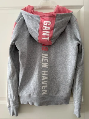 Grå hoodie med dragkedja från Gant - Hoodie från Gant med dragkedja och rosa och vita detaljer. Stl. 158/164 (13-14 år). Köparen betalar frakt. 🩷🤍