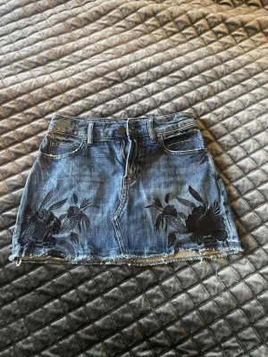 Blå jeanskjol med blommor från Guess - Snygg blå jeanskjol från Guess Äkta med svarta blommiga broderier framtill. Kjolen har rå kant nertill, klassiska fickor fram och bak samt bälteshällor. Perfekt för en trendig och avslappnad look. midjemått ca 35 plus lite stretch