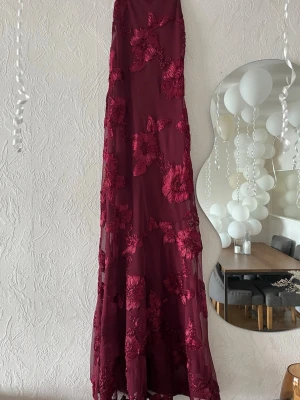 Vinröd långklänning med blommor - Elegant vinröd långklänning med blommiga broderier och transparenta partier. Klänningen har en figurnära passform och är perfekt för speciella tillfällen. Materialet känns lätt och faller snyggt längs kroppen.