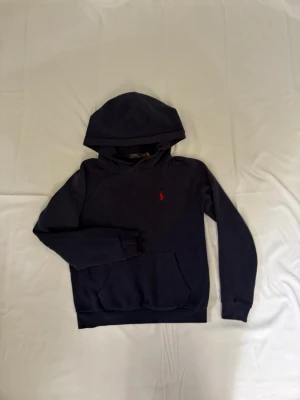Mörkblå hoodie från Ralph Lauren - Mörkblå hoodie från Ralph Lauren med klassisk huva och känguruficka. På bröstet finns den ikoniska röda Polo-loggan broderad. Hoodien har långa ärmar och ribbade muddar vid ärmslut och nederkant. Perfekt för en avslappnad och stilren look.
