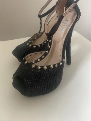 Svarta pumps med nitar och strass - Snygga svarta pumps med höga klackar, öppen tå och T-rem. Skorna är dekorerade med små svarta strasstenar och silverfärgade nitar längs kanterna. Materialet är textil med syntetdetaljer och spänne vid ankeln.