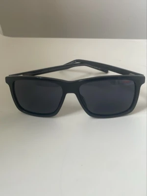 Svarta solglasögon från Hugo Boss - Snygga svarta solglasögon från Hugo Boss med klassisk rektangulär form och mörka glas. Bågarna är i plast och har en stilren design med diskret logga på sidan. Perfekta för dig som vill ha en clean och tidlös look.