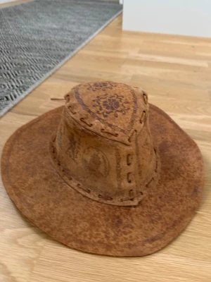 Brun cowboyhatt i mocka - Säljer en brun cowboyhatt i mocka med dekorativa mönster och sömmar runt hela hatten. Hatten har bred brätte och detaljerade tryck på kronan och sidorna. Perfekt för dig som vill sticka ut med en unik accessoar.