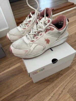 Vita och rosa sneakers från U.S. Polo Assn. - Snygga sneakers i vitt och ljusrosa från U.S. Polo Assn. Skorna har en sportig design med mesh och syntetmaterial, rosa detaljer och logga på sidan. Låga med klassisk snörning och vadderad insida för extra komfort.