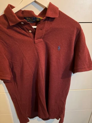 Vinröd pikétröja från Polo Ralph Lauren - Klassisk vinröd pikétröja från Polo Ralph Lauren i custom slim fit. Tröjan har korta ärmar, krage och knappar framtill samt den ikoniska broderade loggan på bröstet. Perfekt för en clean och stilren look.