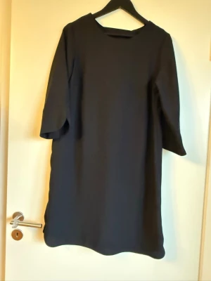Svart enkel klänning med trekvartsärm - Stilren svart klänning med rund halsringning och trekvartsärm. Klänningen har en rak passform och är tillverkad i ett mjukt, något stretchigt material. Ärmarna har en liten slitsdetalj som ger en modern touch.