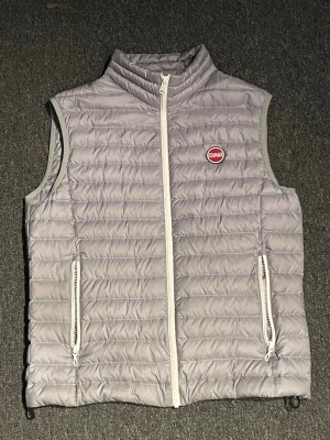 Ljusgrå dunväst från Colmar - Snygg ljusgrå dunväst från Colmar med quiltad design och två dragkedjeförsedda fickor framtill. Västen har hög krage, vit dragkedja och Colmar-logga på bröstet. Perfekt för lager-på-lager och riktigt clean look.