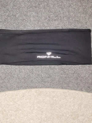 Svart pannband från Ronhill - Säljer ett svart pannband från Ronhill med reflekterande detaljer på baksidan och logga framtill. Tillverkat i stretchigt funktionsmaterial som passar perfekt för löpning eller utomhusaktiviteter. Snygg och enkel design som håller håret på plats.