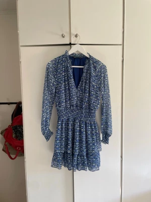 Blå blommig kortklänning från Zara - Supersöt blå kortklänning från Zara med blommigt mönster, v-ringning och långa ärmar. Klänningen har smock i midjan och volangdetaljer vid halsen. Perfekt för dig som gillar en feminin och trendig stil.