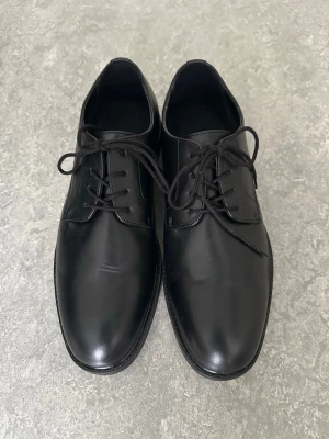 Svarta klassiska loafers i skinn - Säljer ett par stilrena svarta loafers med snörning. Skorna har en slät ovandel i blankt skinn och en klassisk rundad tå. Yttersulan är svart och har en diskret struktur. Perfekta för dig som gillar en clean och tidlös look.