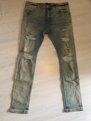 Ljusblå slitna skinny jeans - Säljer ett par ljusblå skinny jeans med flera stora slitningar och hål på båda benen. Jeansen har klassisk femficksmodell, knappgylf och är tillverkade i denim. Perfekta för en trendig och avslappnad stil. ( Amiri jeans) ( tajt i storlek) 