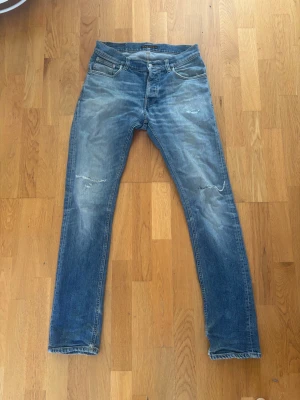 Blå slitna jeans från Nudie Jeans - Säljer ett par blå jeans från Nudie Jeans med  slitningar på både fram- och baksida. Jeansen har klassisk femficksmodell, dragkedjegylf och Nudie Jeans signaturbrodyr på bakfickan. Model: grim Tim 