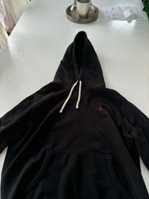Svart hoodie från Polo Ralph Lauren - Svart hoodie från Polo Ralph Lauren med klassisk huva och vita dragsnören. Har en känguruficka framtill och den ikoniska röda loggan broderad på bröstet. Tillverkad i mjukt bomullsmaterial som känns skönt mot huden.