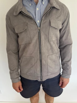 Grå overshirt  - Snygg grå overshirt med fyra fickor framtill och dragkedja. Jackan har klassisk krage och lång ärm, perfekt för dig som vill ha en stilren och modern look. Passar till skjorta eller t-shirt för en avslappnad men uppklädd stil. Kom privat för frågor om overwatch! Aldig använd!