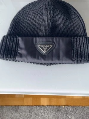 Svart ribbstickad mössa från Prada - Säljer en svart ribbstickad mössa från Prada med bred uppvikt kant och ikonisk trekantig logga framtill i nylon. Mössan är tillverkad i mjukt garn och har en klassisk, tidlös design som passar perfekt till höst och vinter.