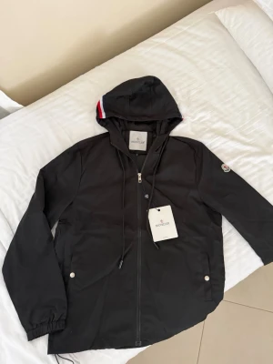 Moncler windbreaker - Snygg svart vindjacka från Moncler med huva och dragkedja framtill. Jackan har två fickor med tryckknappar, elastiska muddar och Moncler-logga på ärmen. Detaljer i rött, vitt och blått på huvan ger en sportig touch. Perfekt för blåsiga dagar.