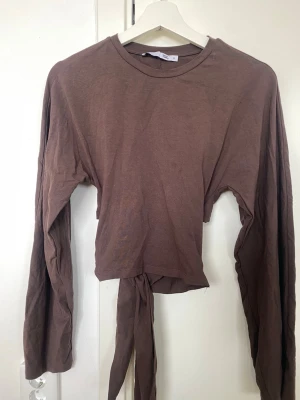 Brun croppad långärmad topp med knyt - Snygg brun croppad topp med långa ärmar och knyt i midjan. Perfekt för dig som gillar en enkel men trendig stil. Tillverkad i mjukt bomullsmaterial och har en rund halsringning.