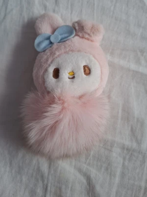 My Melody nyckelring - Nyckelring med My Melody i mjuk rosa plysch och pompom. Ansiktet är broderat och dekorerat med en blå rosett på ena örat. Perfekt att hänga på väskan eller nycklarna för en gullig touch.