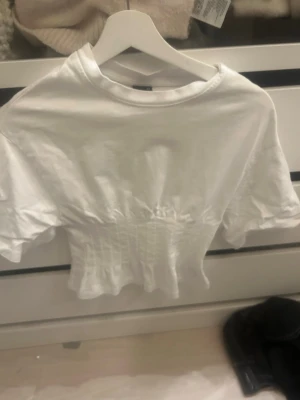 Vit croppad topp från Gina Tricot - Snygg vit croppad topp från Gina Tricot med korta ärmar och smockad midja som ger en figurnära passform. Tillverkad i mjuk bomull och perfekt för en trendig vardagslook.