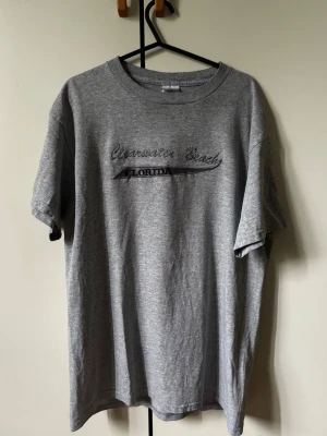Grå Clearwater Beach Florida Broderad  t-shirt - Säljer en grå t-shirt med trycket 'Clearwater Beach Florida' på bröstet. T-shirten har klassisk passform, rund hals och korta ärmar. Perfekt för dig som gillar enkel och avslappnad stil.