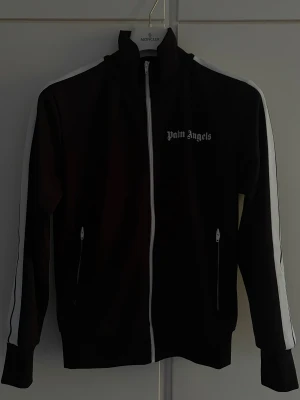 Palm Angels kofta (ÄKTA) - Svart track jacket från Palm Angels med vit dragkedja och vita ränder längs ärmarna. Hög krage och två fickor med dragkedja framtill. Ikonisk logga på bröstet. Äkta och kvitto finns. 