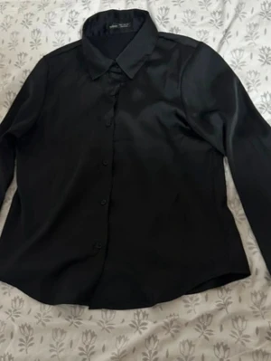 Svart långärmad skjorta i satin - Stilren svart skjorta i glansigt satinmaterial med klassisk krage och knäppning framtill. Skjortan har långa ärmar och en rak passform som passar till många olika looks. Perfekt för dig som gillar en clean och tidlös stil.